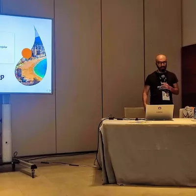 Ricardo Sanz en el estado hablando junto a una pantalla con la diapositiva de presentación de la charla "Testeándolo todo: testing más allá de PHPUnit"